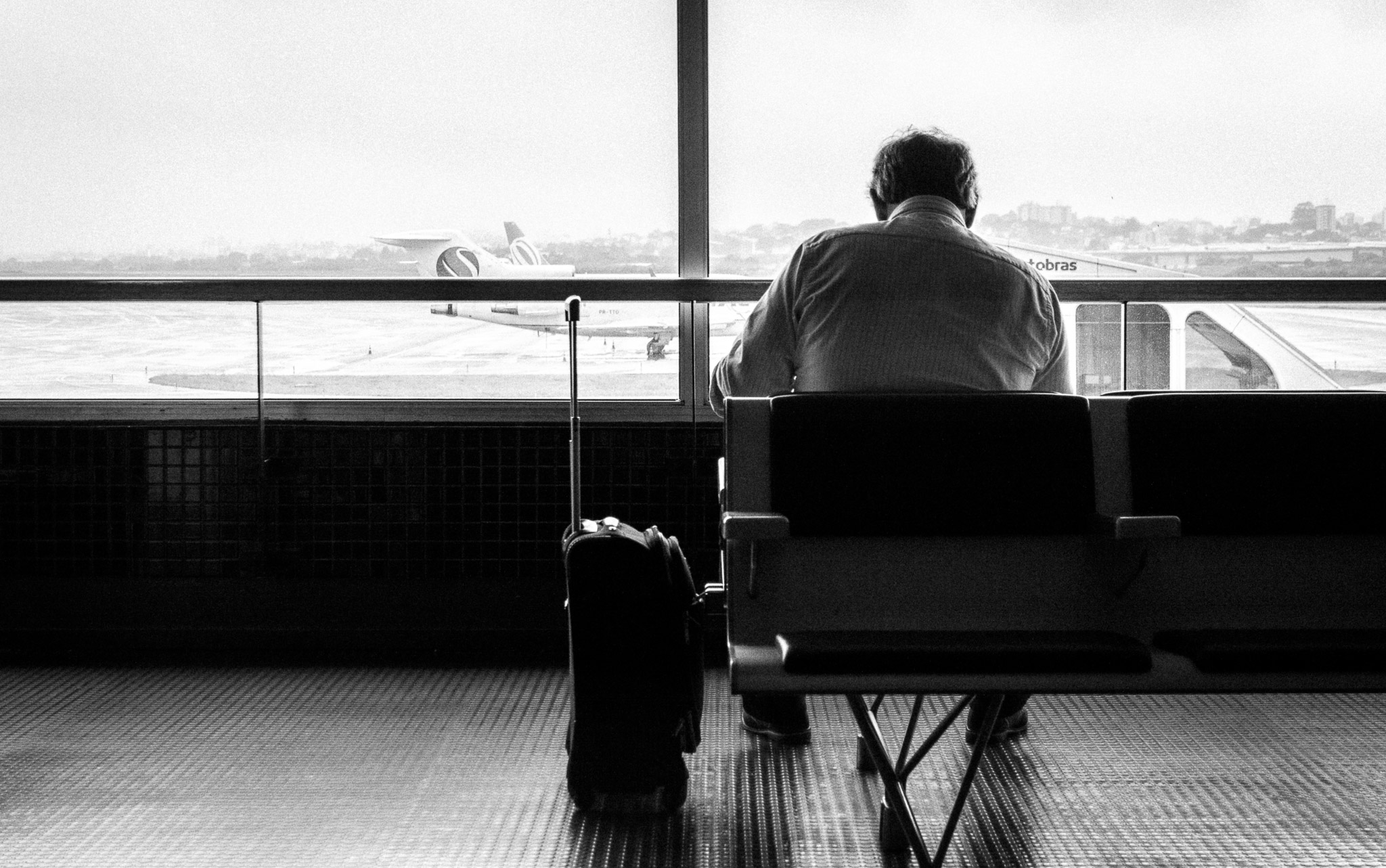 Black and white shot of a man seen from behind in an airport lounge, suitcase beside him, planes on the tarmac