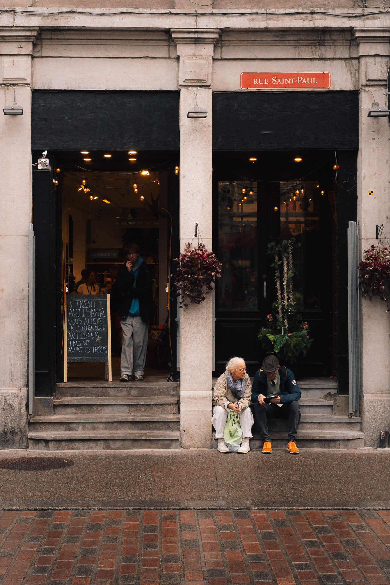 Two older men sitting on the steps of a shop entrance on rue Saint-Paul