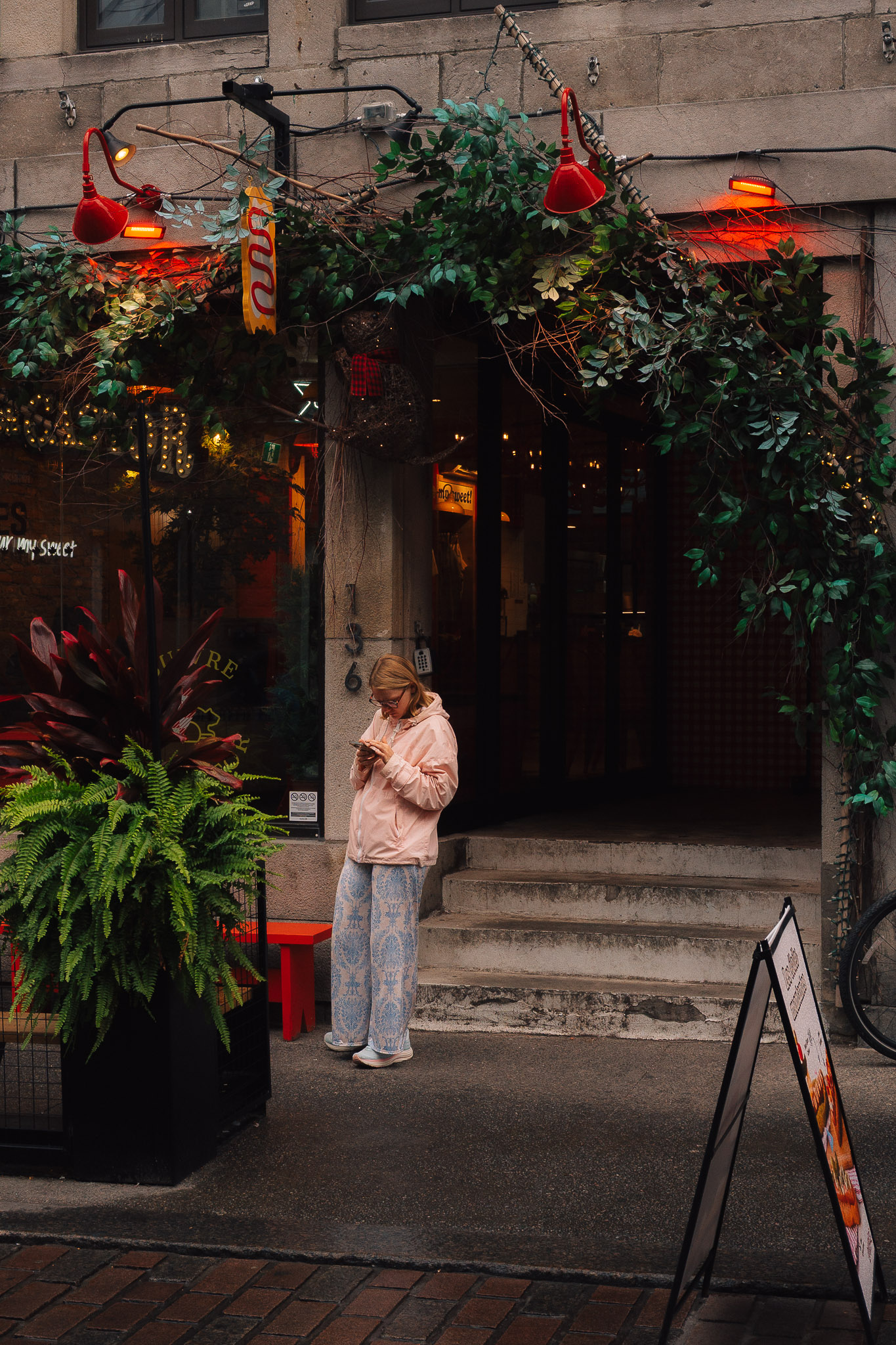 Woman in a pink jacket looking at her phone under a doorway draped in vines