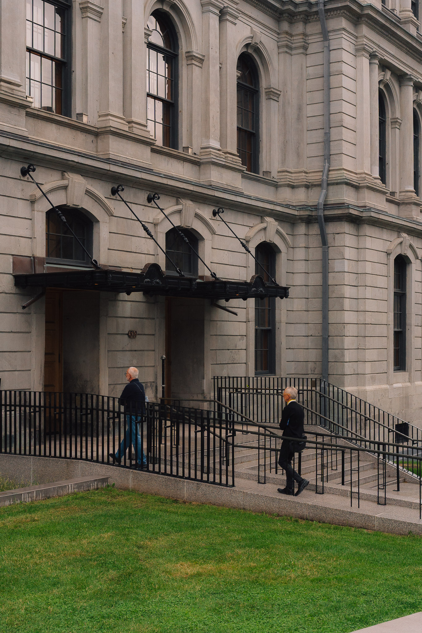Two grey-haired men walking up railed ramps in front of an old stone building