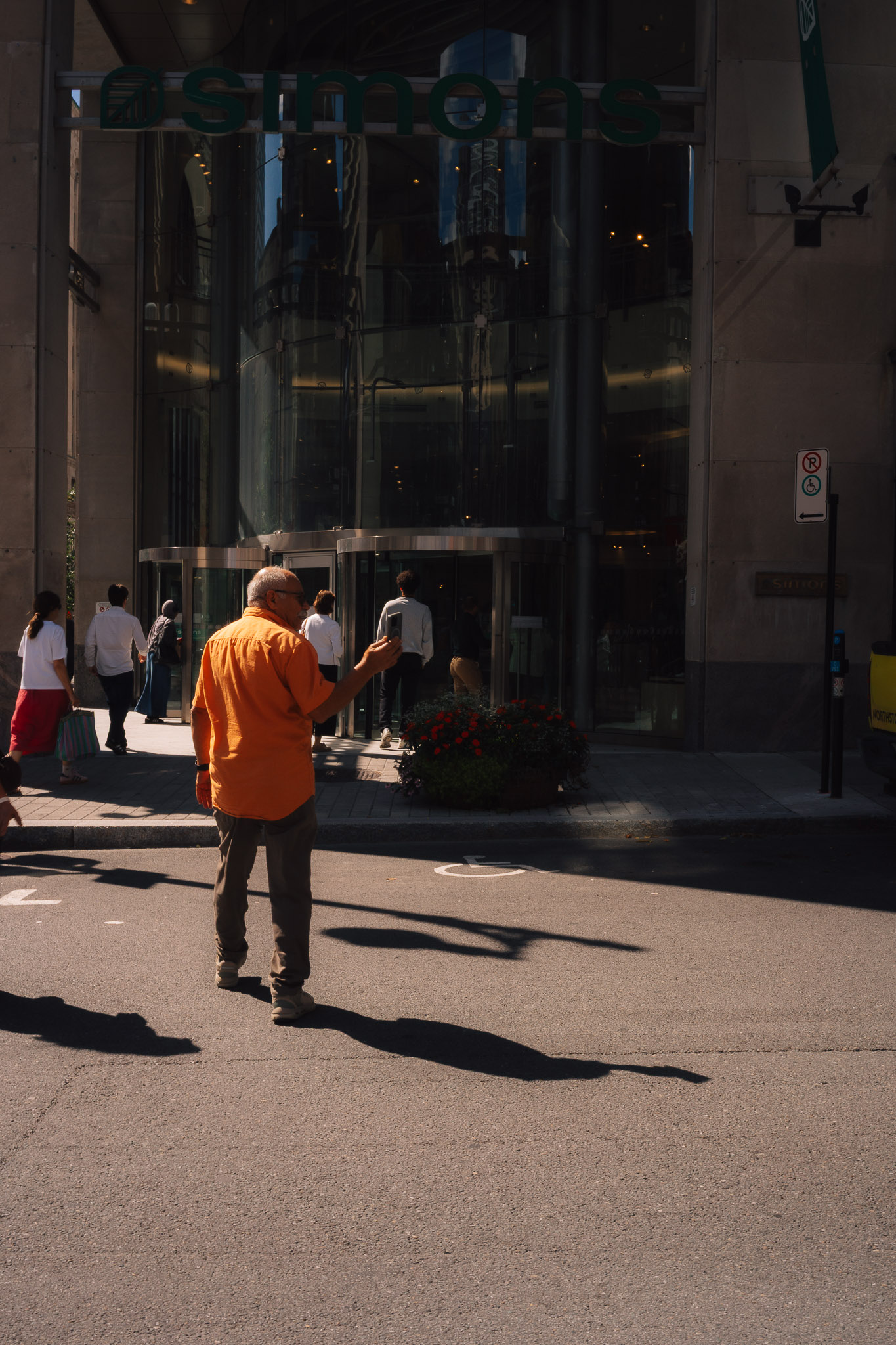 Man in an orange shirt crossing a sunlit street, his long shadow stretching beside him