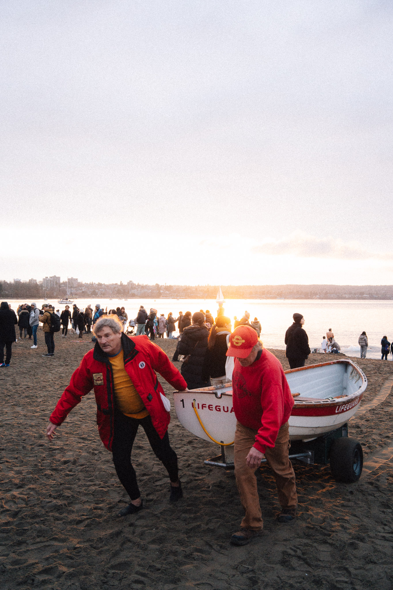 Two lifeguards in red jackets hauling a rescue boat across the sand in front of a crowd