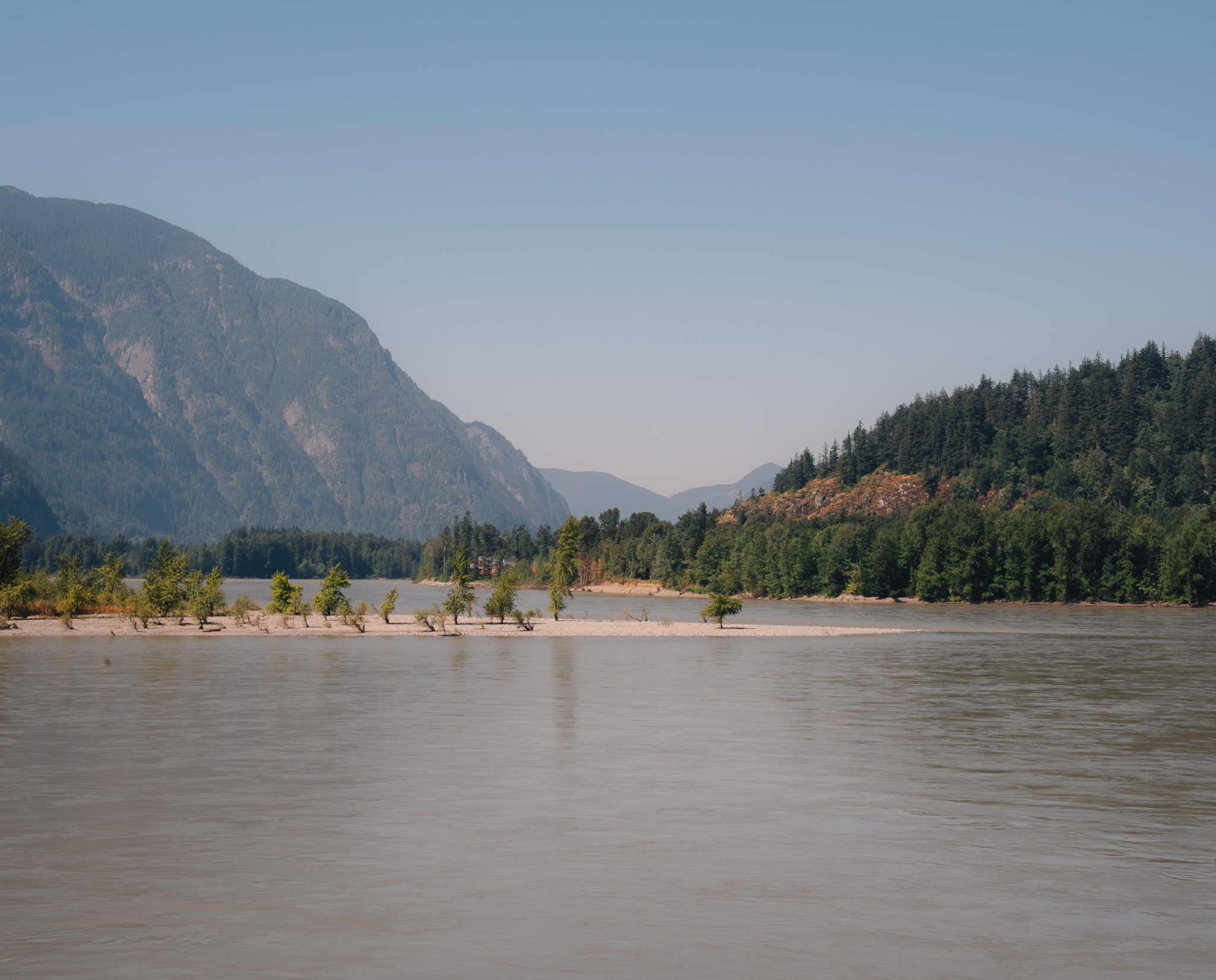 Silt-grey river running past a wooded gravel bar, hazy mountains behind