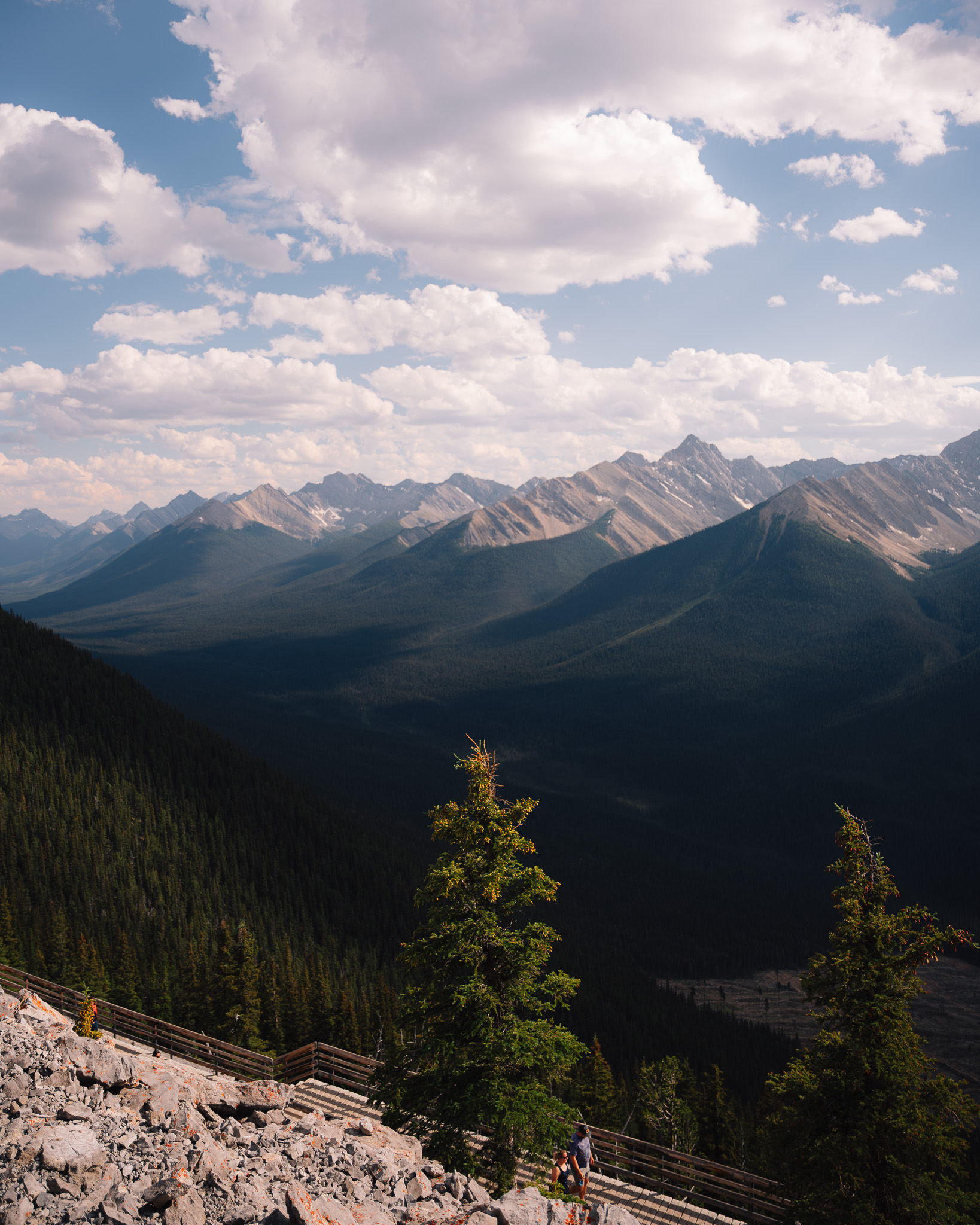 Hiker on a gravel path high above a forested valley ringed by peaks