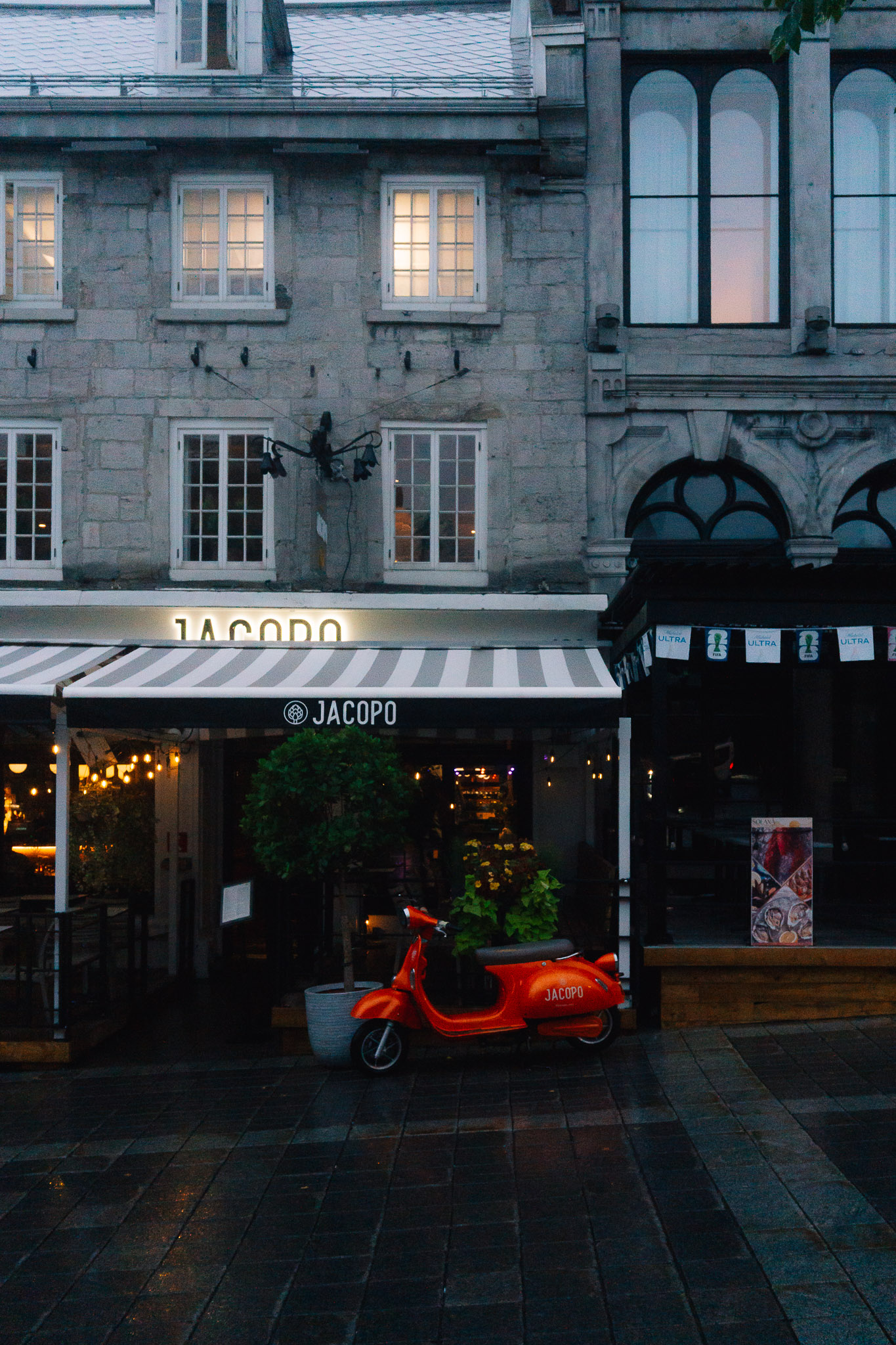 Red scooter parked in front of a restaurant's striped awning on a wet grey street