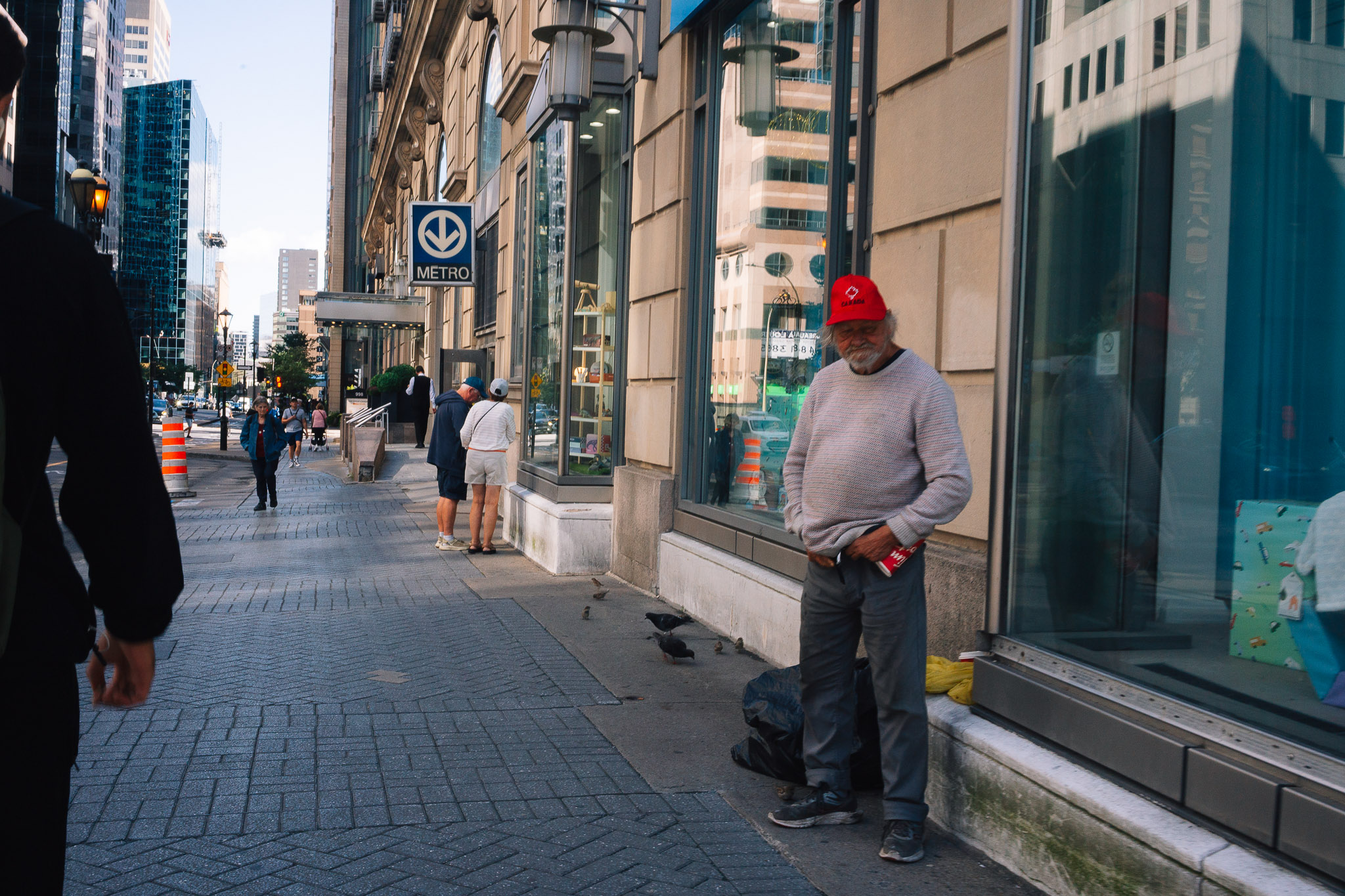 Man in a red cap leaning against a storefront on a downtown sidewalk, pigeons at his feet