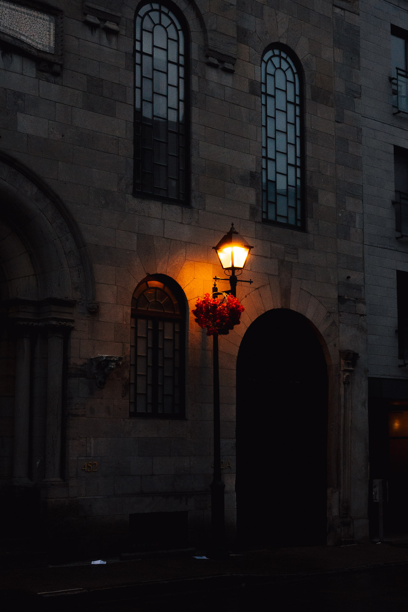 Street lamp glowing at dusk beside a hanging basket of red flowers on a dark stone facade