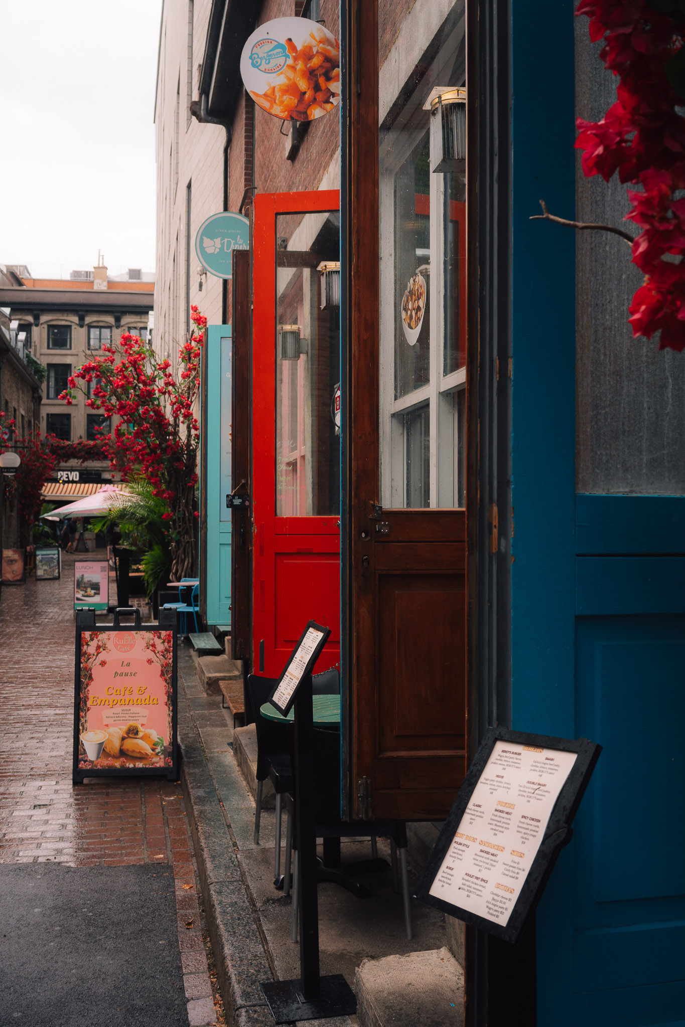 Row of shopfronts on a wet side street, a red door between blue and wooden ones under red flowers